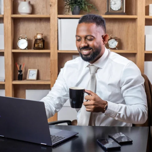 homem negro segurando xicara, em frente ao notebook em um escritorio